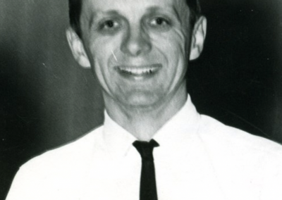 Black and white portrait of Ron Underwood during his early career as a speech and debate coach, smiling warmly in a collared shirt and tie.